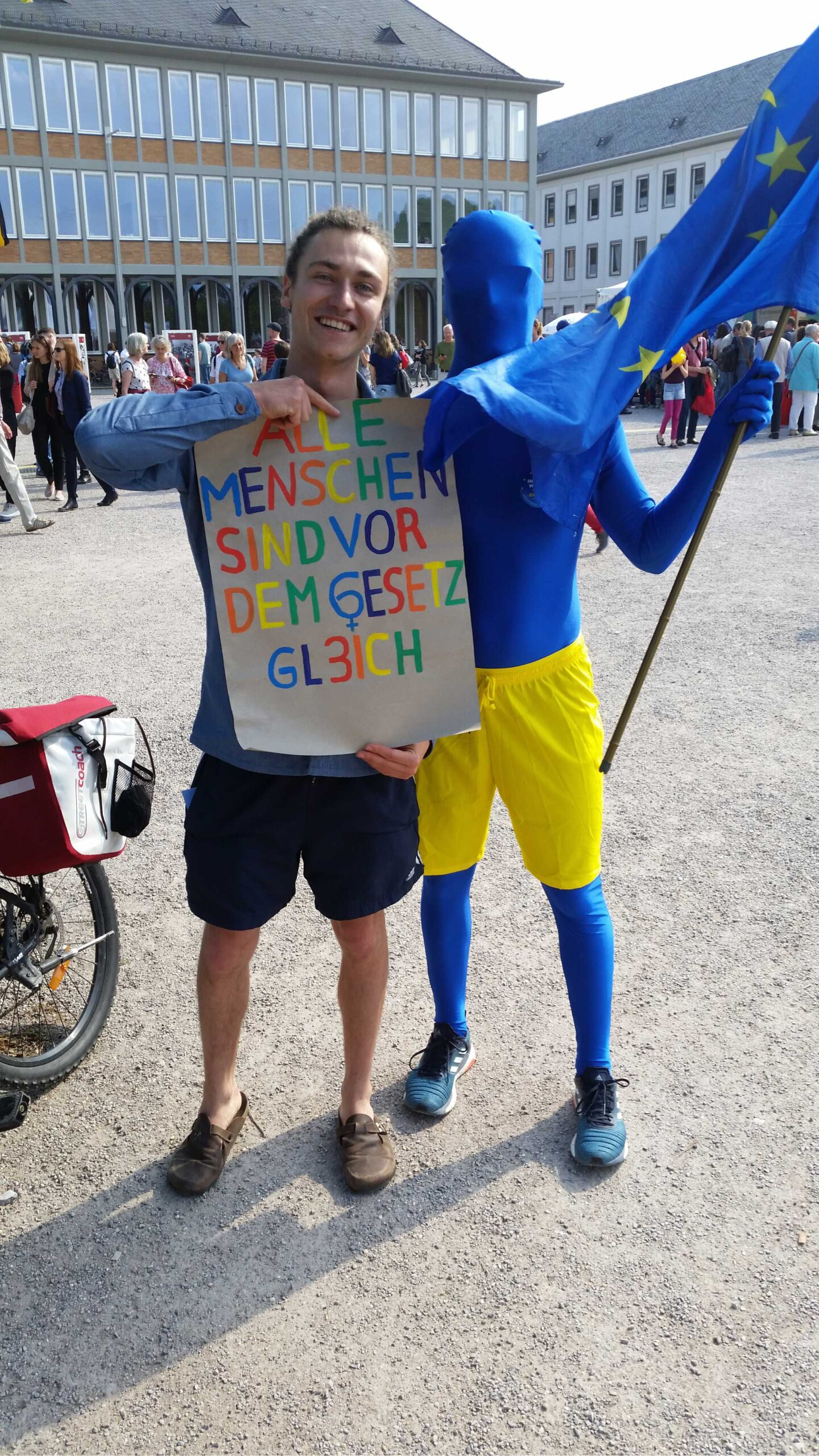 Kulturhunt Karlsruhe: Zeige uns eines der Menschenrechte Ein junger Mann steht mit einem Plakat "Alle Menschen sind vor dem Gesetz gleich" in der Stadt, direkt neben ihm eine als Europa verkleidete Person in blauem Ganzkörperanzug und mit Europa-Flagge