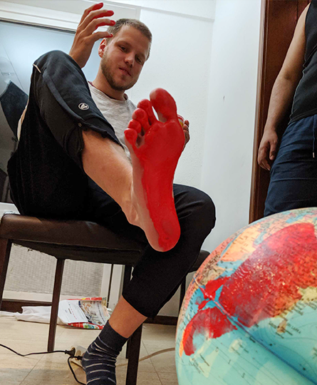 Junger Mann sitzt vor einem Globus auf einem Stuhl, einen Socken hat er ausgezogen und die Fußsohle ist bedeckt mit roter Farbe. Auf dem Globus ist entsprechend ein roter Fußabdruck