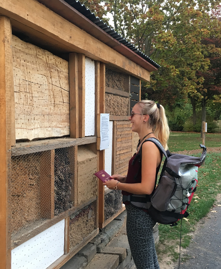 Eine Frau mit Rucksack und Reisepass in der Hand steht lachend vor einer großen Insektenbehausung