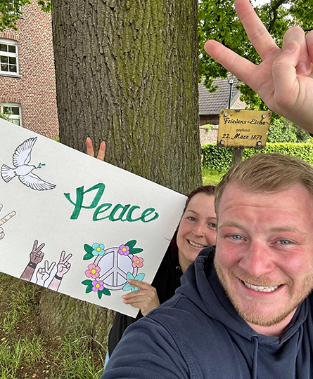 Zwei Personen halten ein Plakat mit Friedenssymbolen hoch und machen dazu das Peace-Zeichen mit ihren Fingern - im Hintergrund steht eine Friedenseiche