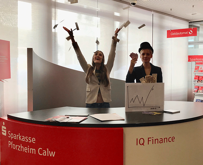 Zwei Personen hinter einem Sparkassen-Tresen, die mit Geld werfen und eine Börsengrafik vor sich stehen haben