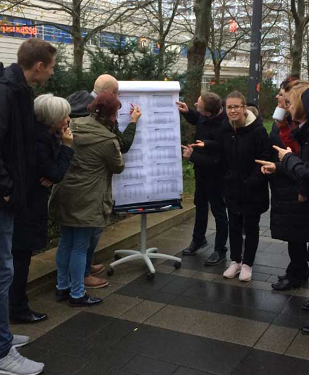 Ein Team aus verschiedenen Menschen stehen draußen auf der Straße um einen rollbaren Flipchart herum, einer hat sogar Kaffeebecher dabei. sie scheinen ein Meeting abzuhalten