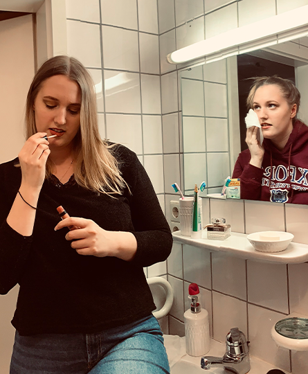 Eine junge Frau in einem Badezimmer trägt sich zur Kamera gerichtet Lipgloss auf. Der Spiegel zeigt eine andere Frau beim Abschminken.