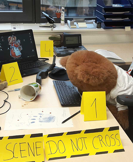 Das Bild zeigt einen Arbeitsplatz als Tatort umdekoriert. Ein Teddy mit Messer im Rücken liegt halb auf der Tastatur, die Beweisstücke wurden mit gelben Zahlen markiert, vorne steht Crime scene, do not cross als Absperrung