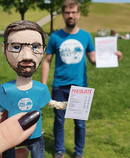 Das Bild zeigt eine detailreich modellierte Puppe mit blauem Shirt und Preisliste in der Hand, verschwommen im Hintergrund ist der Kollege zu erkennen, der eben dieses Shirt trägt und auch eine Preisliste in der Hand hält