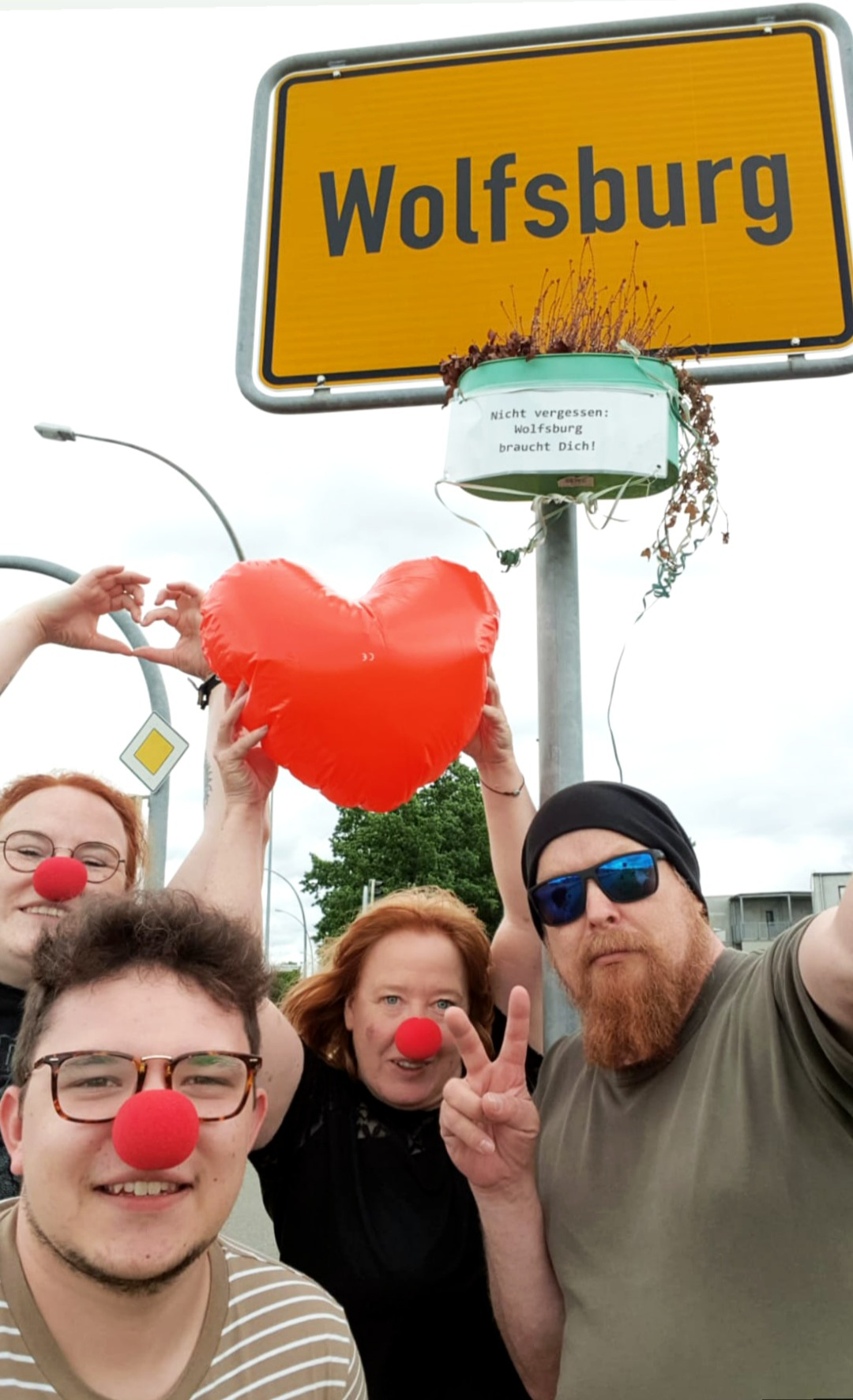 Scavenger Hunt Wolfburg: viel Liebe für die Stadt Unter einem Ortsschild von Wolfsburg stehen vier Menschen mit einem Herz, drei Clownsnasen und einer zeigt das Peace-Zeichen