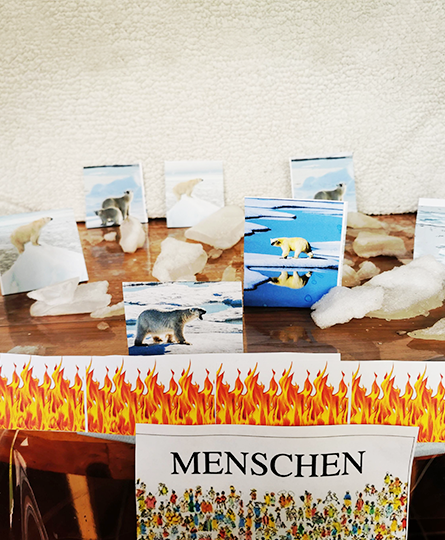Auf einem Tisch drapiert: Bilder von Eisbären zwischen schmelzenden Eis-Brocken, davor angedeutetes Feuer und Menschen, die dieses verursachen