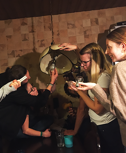 Fünf Personen haben sich um eine Deckenlampe versammelt, reichen Glühbirnen an und schauen skeptisch, während zwei scheinbar mühsam die alte Birne entfernen