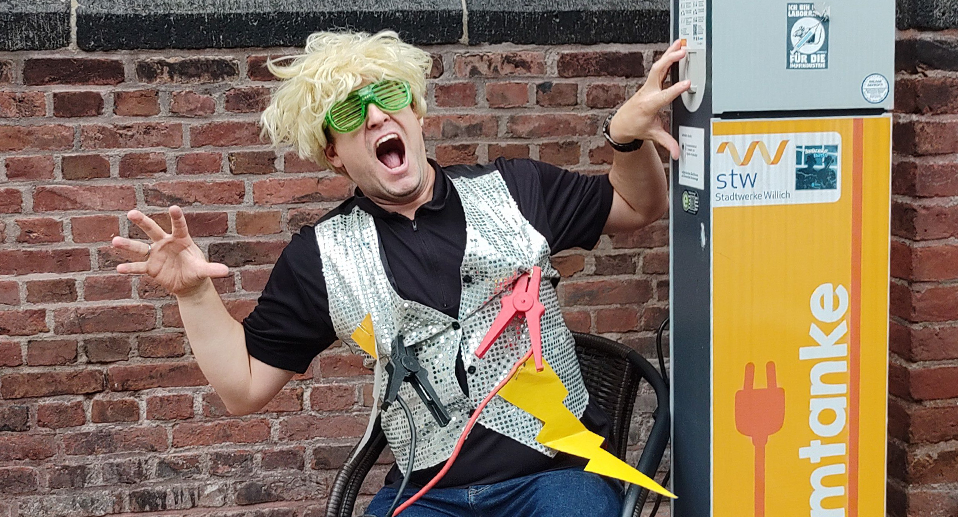 Teammitglied nutzt eine E-Mobilität Tanksäule, um sich wieder aufzuladen, mit Perücke und aufgeklebten Blitzen