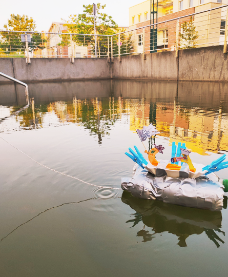 Ein Boot aus Plastikflaschen, die mit Klebeband verbunden wurden, treibt auf einem städtischen Gewässer. An Bord kleine Figuren und Wäscheklammern.