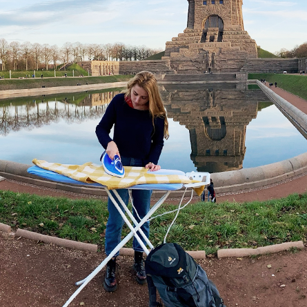 Junge Frau bügelt mitten in einem Park vor einer Sehenswürdigkeit, deuter Rucksack unten im Bild