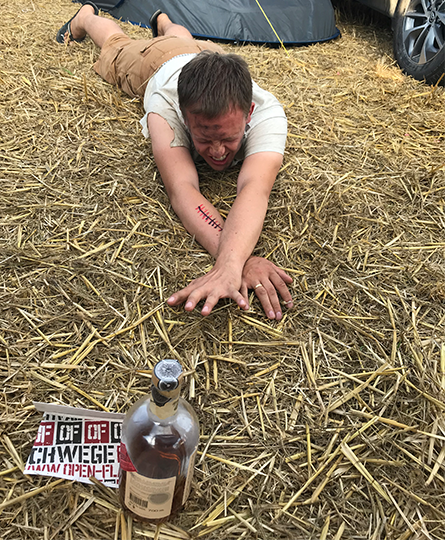 Ein Teilnehmer robbt flach über das Stroh am Boden und reckt sich nach einem Festivalticket und einer Flasche Alkohol