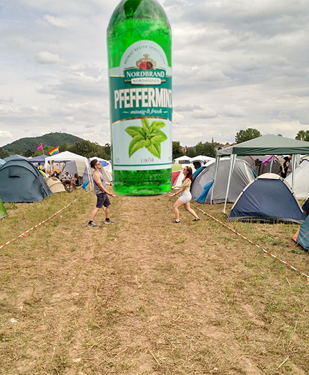 Zwei Personen auf einem Zeltplatz, die gemeinsam zwischen sich eine scheinbar überdimensionale Pfefferminzlikör-Flasche hochhalten - eine gelungene Illusion