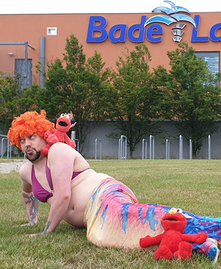 Vor dem Freizeitbad "Badeland" in Wolfsburg liegt ein als Nixe verkleideter Mann auf dem Rasen und posiert mit witzigem Gesichtsausdruck.