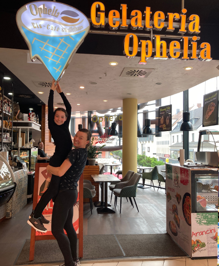 Eine junge Mutter hebt ihre Tochter hoch, damit es so aussieht als hielte diese die Eiswaffeln aus dem Gelateria Ophelia Schriftzug in der Hand.
