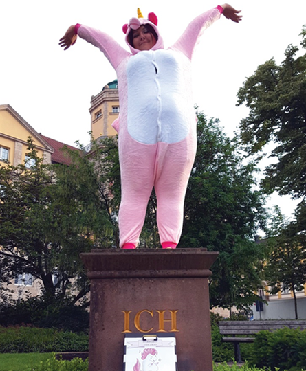 Auf einem Podest mit der Aufschrift "ICH" steht eine als Einhorn verkleidete Dame und streckt die Hände hoch