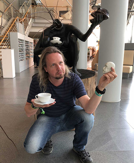 Mann mit längeren Haaren hockt vor einer Skulptur und imitiert diese. Dazu hält er eine Teetasse und einen Kopf in der Hand, die Skulptur ist ebenfalls mit Tasse und Schädel.