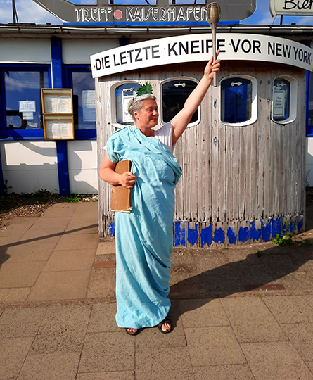 Eine Dame steht als Freiheitsstatur verkleidet vor der Kneipe mit dem Namen "Die letzte Kneipe vor New York".
