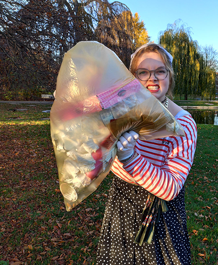 Eine junge Frau hält in einem Park einen Sack mit Müll hoch, den sie gesammelt hat - dabei schneidet sie eine grimmige Grimasse