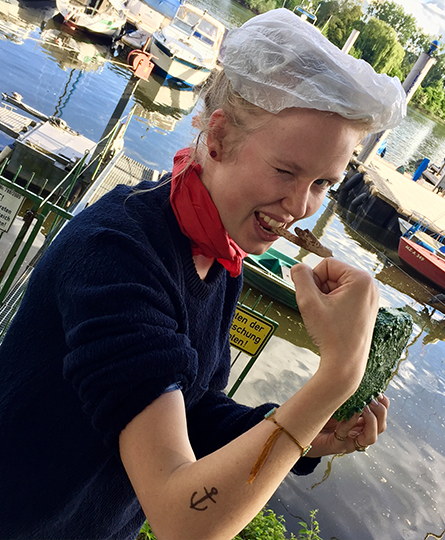 Junge Frau als Popeye verkleidet: sie flext den Arm mit einer Faust, in der anderen Hand hält sie gefrorenen Spinat, im Mund ein Stock als Pfeife und als Mütze dient eine Mülltüte. Sie zwickert in die Kamera