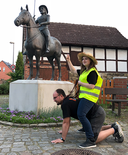 Das Bild zeigt ein Reiterdenkmal, davor ein junger Mann auf Händen und Knien, also als Pferd, und eine junge Frau mit Strohhut, die auf ihm sitzt, als würde sie reiten