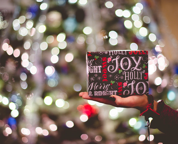Eine Hand hält ein Geschenk hoch mit dem Wort Joy, im Hintergrund ein beleuchteter Weihnachtsbaum